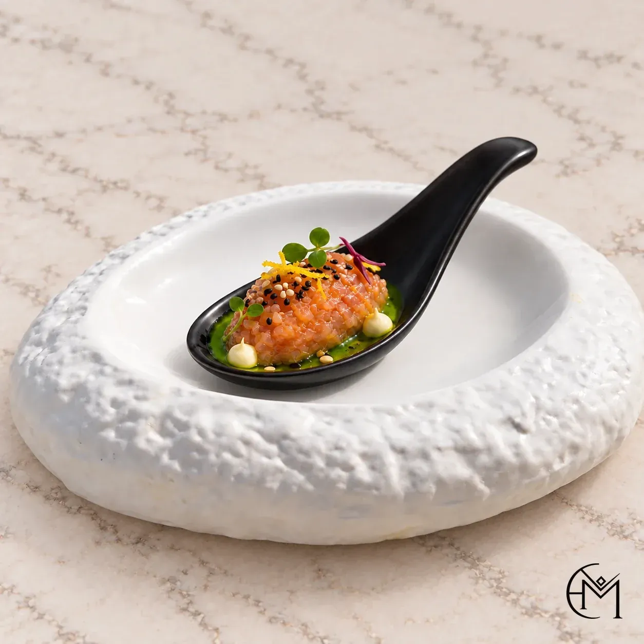 A spoonful of tartare topped with sesame, lemon zest and micro herbs, on herb oil in a black spoon, set in a textured white bowl.